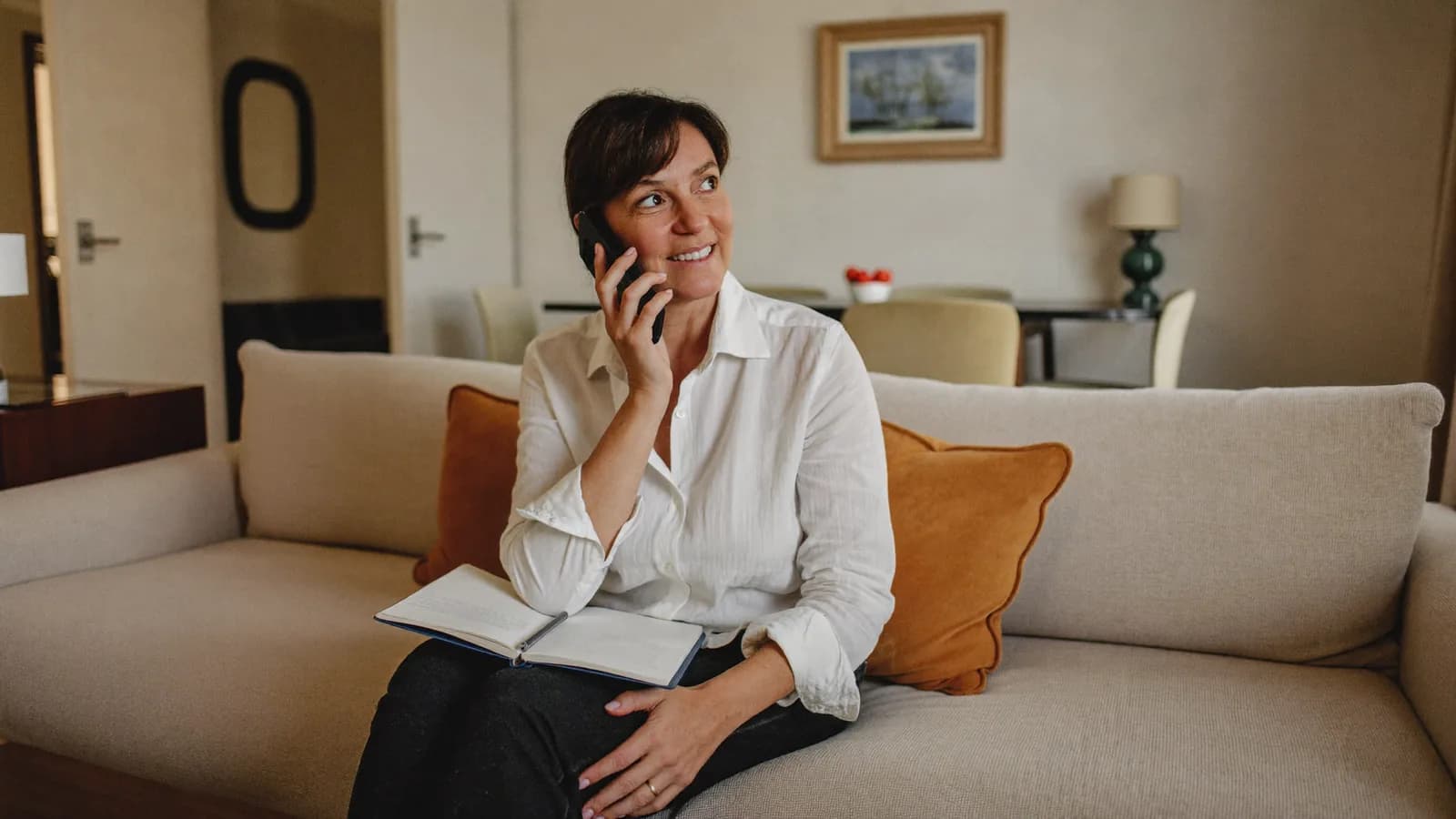 Mujer sentada en el sofá de su casa hablando por teléfono con una libreta en el regazo